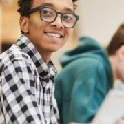 Cheerful excited smart black student in glasses sitting at table and looking at camera while using laptop in library