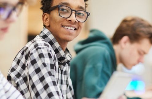 Cheerful excited smart black student in glasses sitting at table and looking at camera while using laptop in library