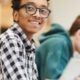 Cheerful excited smart black student in glasses sitting at table and looking at camera while using laptop in library