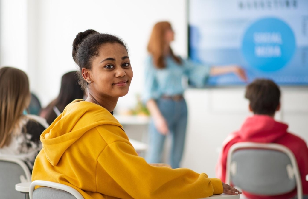 Happy college student sitting in school-desk and looking in camera.