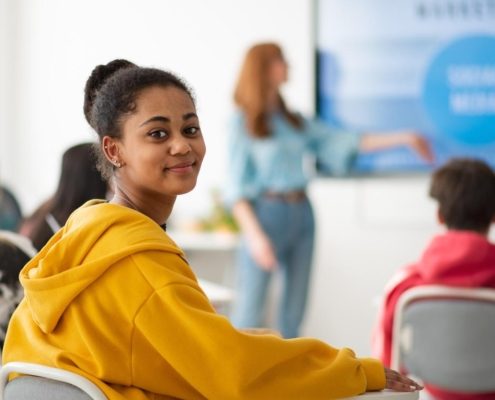 Happy college student sitting in school-desk and looking in camera.