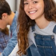 Satisfied young woman looking at camera while her friends studying at college. Team of multiethnic students preparing for university exam. Portrait of beautiful girl with freckles sitting in a row with her classmates during high school exam.