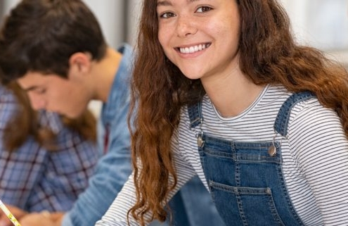 Satisfied young woman looking at camera while her friends studying at college. Team of multiethnic students preparing for university exam. Portrait of beautiful girl with freckles sitting in a row with her classmates during high school exam.