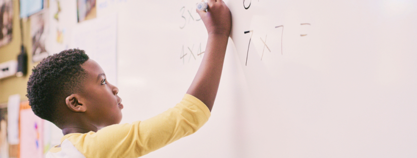 A young black boy enthusiastically writing a math problem on a whiteboard, fully engaged in his learning process.