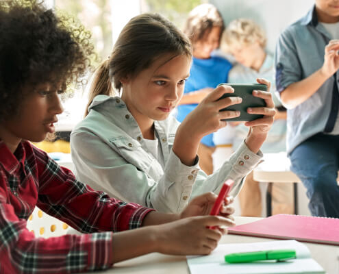 Middle school age female student sits at a table holding her smartphone in front of her instead of doing her school work. Another student sits next to her and students can also be seen behind her.