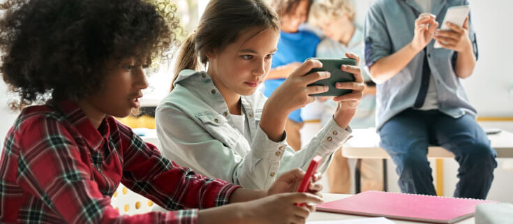 Middle school age female student sits at a table holding her smartphone in front of her instead of doing her school work. Another student sits next to her and students can also be seen behind her.