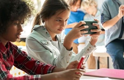 Middle school age female student sits at a table holding her smartphone in front of her instead of doing her school work. Another student sits next to her and students can also be seen behind her.