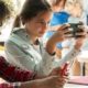 Middle school age female student sits at a table holding her smartphone in front of her instead of doing her school work. Another student sits next to her and students can also be seen behind her.