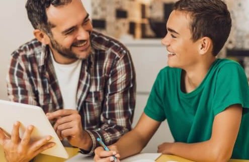 Father and son sit at the kitchen table with the son's homework spread out in front of them. Both are smiling.