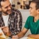 Father and son sit at the kitchen table with the son's homework spread out in front of them. Both are smiling.