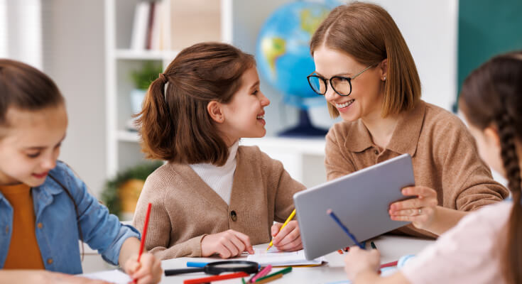 Female adult with brown hair and glasses smiles at an elementary school age girl student who smiles back. The teacher holds a tablet in front of both of them and another student works quietly to the side.