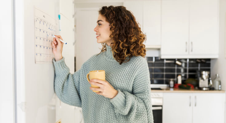 Parent marking something on a hanging family calendar while holding a coffee.