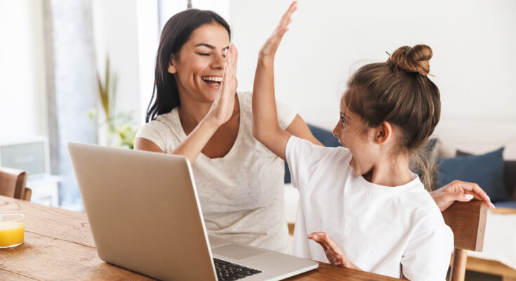 Mom and young daughter high five while sitting in front of an open laptop.