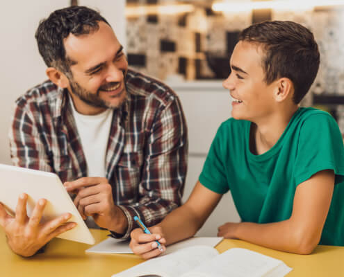 Father and son sit at the kitchen table with the son's homework spread out in front of them. Both are smiling.