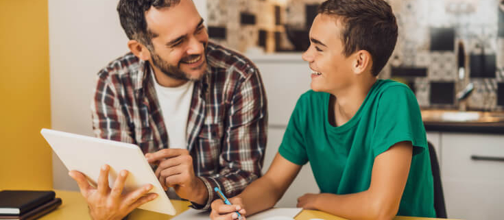 Father and son sit at the kitchen table with the son's homework spread out in front of them. Both are smiling.