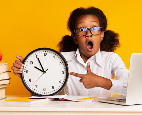Young Black girl student is holding and pointing to an analog clock with her mouth open. The background is yellow and there are school supplies on the table.