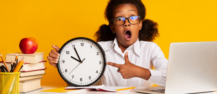 Young Black girl student is holding and pointing to an analog clock with her mouth open. The background is yellow and there are school supplies on the table.