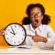 Young Black girl student is holding and pointing to an analog clock with her mouth open. The background is yellow and there are school supplies on the table.