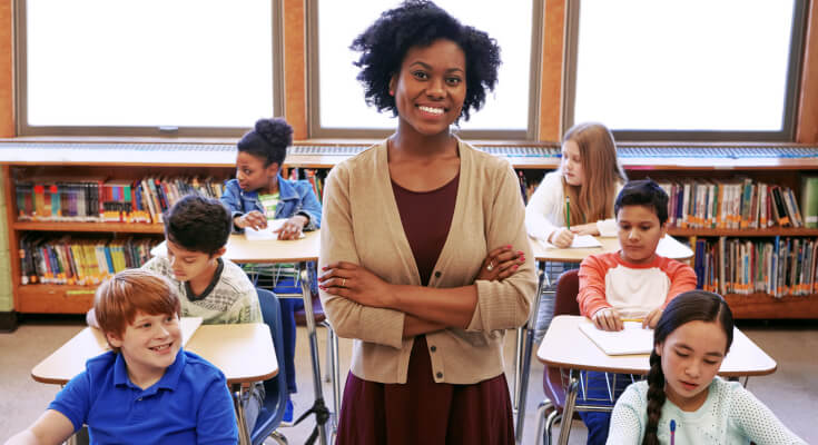 A young female Black teacher stands smiling at the camera with students sitting at desks behind her.