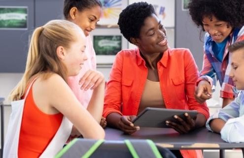 A group of four young students are gathered around a female teacher who smiles and shows them a lesson on a tablet.