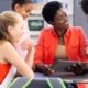 A group of four young students are gathered around a female teacher who smiles and shows them a lesson on a tablet.