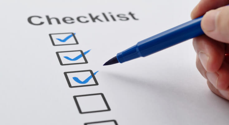 Close up view of a hand holding a blue pen over a piece of paper. The paper shows the words "Checklist" and three checked boxes and one unchecked box.