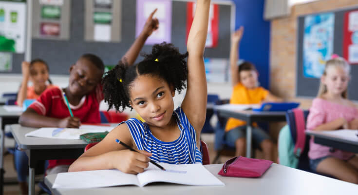 Elementary school female student raises her hand during class, with other students raising their hands in the background. She holds a pencil over an open workbook.