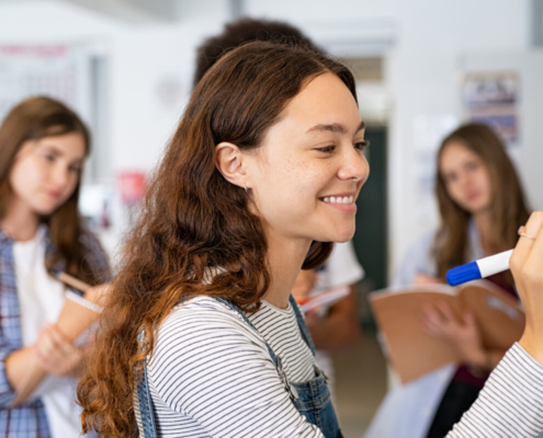 Female teenage high school student with long brown hair smiles as she writes on a white board. Other students look on in the background.