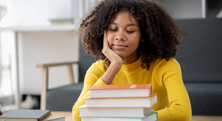 Female high school student sits with her head propped up by her hand, looking glumly at a stack of books sitting in front of her.