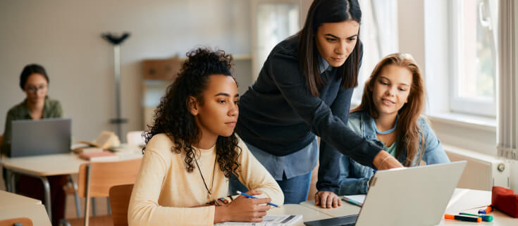 A teacher leans over a desk where two female high school students sit and review something on an open laptop.