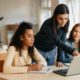 A teacher leans over a desk where two female high school students sit and review something on an open laptop.