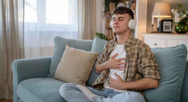Male teenager sits on a couch with legs crossed and over-the-ear headphones on. He has his eyes closed and one hands on his chest as he meditates.