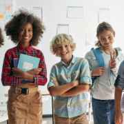 Diverse group of five middle school aged students who stand in front of a window inside a school. The students are smiling and holding various school supplies, like books and backpacks.