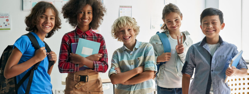 Diverse group of five middle school aged students who stand in front of a window inside a school. The students are smiling and holding various school supplies, like books and backpacks.