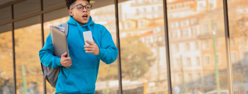 Middle school boy in blue sweatshirt and holding a backpack is running in front of the school as if he's late