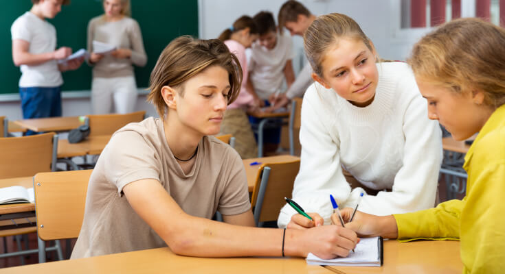 Three middle school students sit at a table with other students in the background. They are writing on the same notebook as they study together.