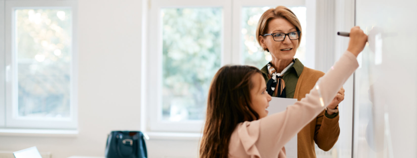 Educator stands next to a white board while an elementary school female student reaches up to write on the whiteboard.