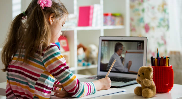Elementary school girl sitting at a laptop while she is being academic coached via video chat. She holds a pencil and is writing notes.