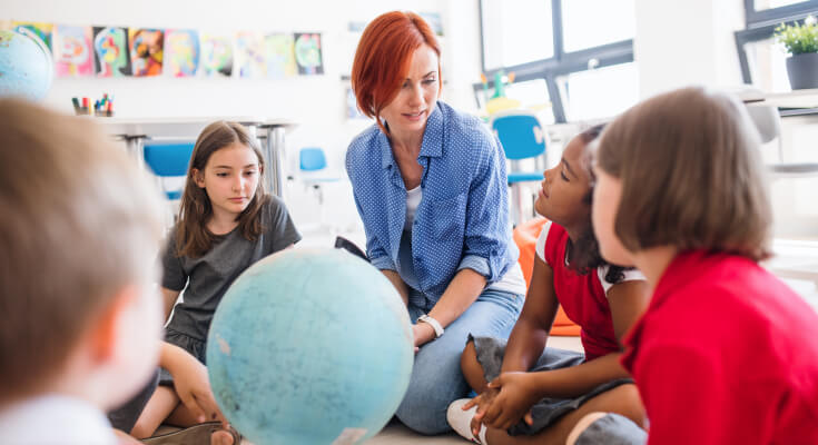 Female academic tutor sits on the floor of a classroom with several elementary school students, having a discussion.
