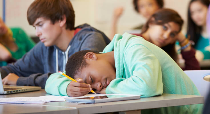Male middle school student in a light green sweatshirt is asleep on his desk, with other students awake and working behind him.