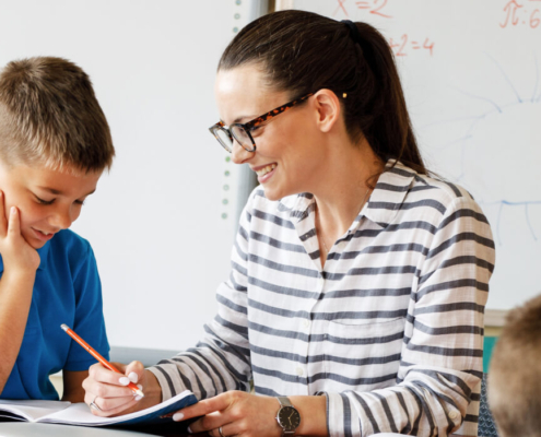 Female teacher with glasses on sits with an elementary school boy student in a blue polo shirt. The boy has his hand on his chin as he looks at something the teacher points out on a notebook in front of them.