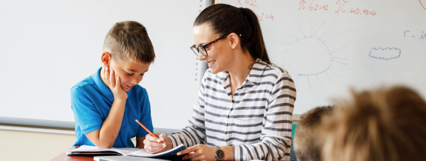 Female teacher with glasses on sits with an elementary school boy student in a blue polo shirt. The boy has his hand on his chin as he looks at something the teacher points out on a notebook in front of them.