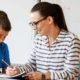 Female teacher with glasses on sits with an elementary school boy student in a blue polo shirt. The boy has his hand on his chin as he looks at something the teacher points out on a notebook in front of them.