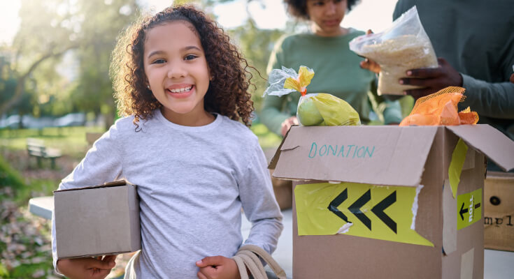 Elementary school age female student stands by a box labeled "donation" during a sunny summer day. She is holding a smaller box and smiling while engaging in a volunteer event.