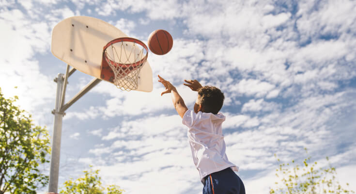 A young boy in a white t-shirt shoots a basketball at a basket. The ball is in the air near the rim and a blue sky with white clouds is behind it.
