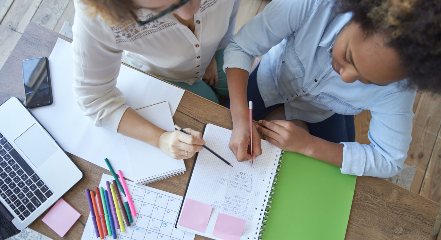 A woman in glasses leans over an elementary school child, who is writing in a notebook. Both are holding pencils. The desk they are sitting at is covered in spiral-bound notebooks, colored markers, a computer, and a calendar.