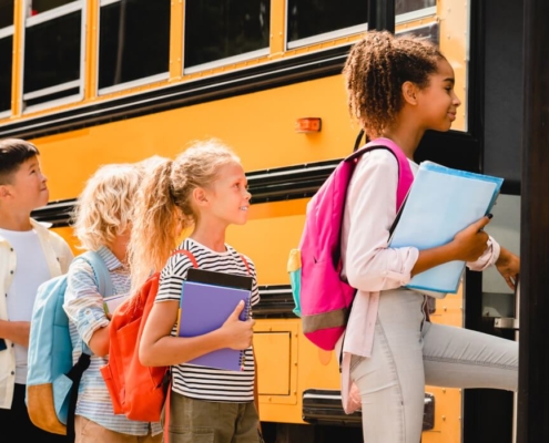 A group of elementary-aged students are standing in line waiting to board a yellow school bus. They are wearing backpacks, holding binders and folders, and look excited.