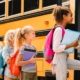 A group of elementary-aged students are standing in line waiting to board a yellow school bus. They are wearing backpacks, holding binders and folders, and look excited.