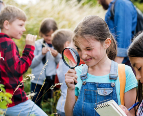 Elementary school class explores outside in a forested area. The focus is on a young girl in a blue shirt and overalls who holds a magnifying glass up to her eye in front of a plant.
