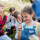 Elementary school class explores outside in a forested area. The focus is on a young girl in a blue shirt and overalls who holds a magnifying glass up to her eye in front of a plant.